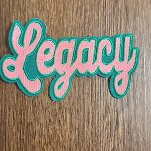 May include: A pink and green embroidered patch with the word "Legacy" in a cursive font. The word is outlined in green and has a pink fill. The patch is on a wooden surface.