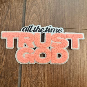 May include: A decorative patch featuring the text all the time in black script lettering above the words TRUST GOD in large, textured, coral-colored block letters. The entire phrase is outlined with a shimmering silver border.