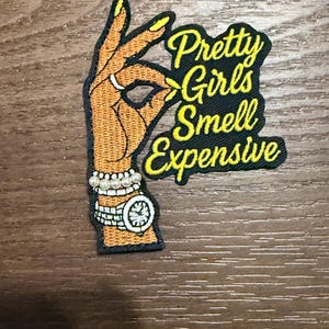 May include: Embroidered patch featuring a hand gesture with a ring and a watch, along with the text "Pretty Girls Smell Expensive" in yellow and black. The patch has a black border and is on a brown surface.