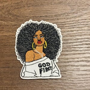 May include: Embroidered patch featuring a Black woman with large, curly black hair and gold hoop earrings. She wears a white top with the words "GOD FIRST" in black lettering. The patch has a white border and is set against a wood grain background.