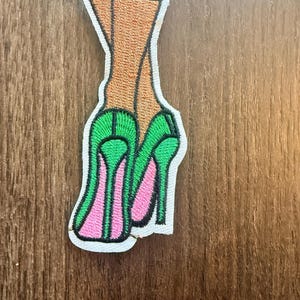 May include: An embroidered patch featuring a pair of legs in tan stockings wearing bright green high-heeled shoes with pink inner soles and black outlines, all set against a white fabric border with a visible stitched edge.