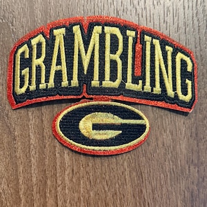 May include: An embroidered patch featuring the word GRAMBLING in gold lettering with a black outline, arched over a smaller oval patch containing a gold letter G on a black background. Both pieces have a shimmering red border trim.