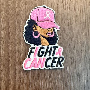 May include: An embroidered patch featuring a person with dark curly hair wearing a pink baseball cap with a white ribbon symbol. The person has pink hoop earrings and pink lipstick. Below, the text reads FIGHT CANCER in black and pink letters.