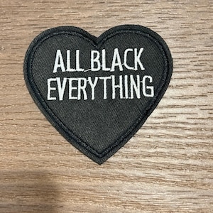 May include: A black heart-shaped patch with the phrase "ALL BLACK EVERYTHING" embroidered in white. The patch has a black border and is likely made of fabric. The design is simple and bold, suitable for clothing or accessories.