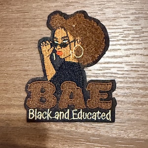 Puede incluir: Parche bordado que representa a una mujer negra con gafas, pendientes de aro dorados y un afro marrón. El parche dice "BAE Black and Educated" en letras marrones sobre un fondo negro. El parche es rectangular con un borde marrón.