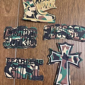 May include: A collection of embroidered patches with a camouflage pattern. The patches include text such as "Prayer Warrior," "Fashion Junkie," "Blessed," and "Fearless Girl." One patch features a high-heeled shoe.