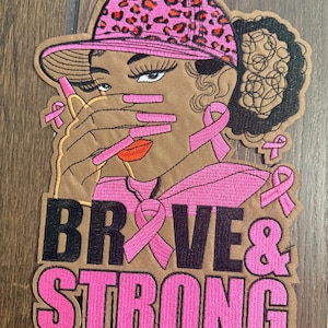 May include: An embroidered patch featuring a person wearing a pink leopard print cap, with long pink nails covering their mouth. The design includes pink awareness ribbons and the text BRAVE & STRONG in black and pink lettering on a tan background.