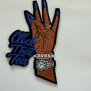 May include: Embroidered patch featuring a brown hand with blue fingernails, a silver ring, and a pearl bracelet. A watch with a white face and silver band is also visible. The patch has the text "Clock That Tea" in blue.
