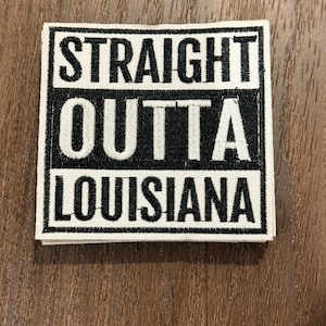 May include: A square patch with the words "STRAIGHT OUTTA LOUISIANA" in bold, black capital letters. The text is on a white background, framed by a black border. The patch is on a wooden surface.