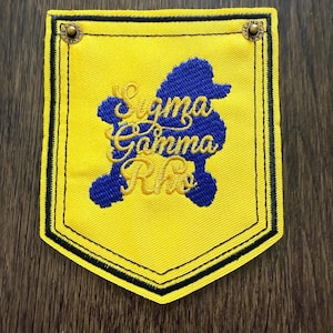 May include: A yellow embroidered patch with a black border and the words "Sigma Gamma Rho" in blue and gold. The patch features a blue poodle design and is secured with two brass-colored rivets.