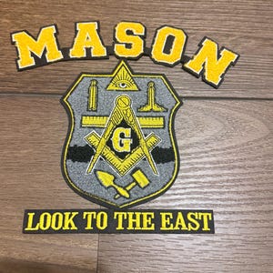 Könnte beinhalten: Ein Freimaurer-Emblem-Patch mit dem Wort "MASON" in gelben Buchstaben über einem grau-gelben schildförmigen Patch. Der Patch zeigt freimaurerische Symbole und den Text "LOOK TO THE EAST" darunter.