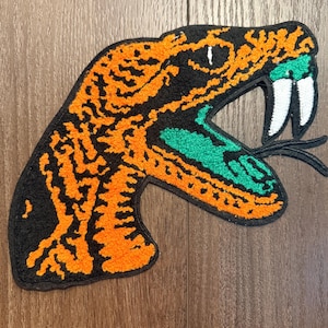 Puede incluir: Un parche textil con forma de cabeza de serpiente de perfil, con una base negra y patrones texturizados de color naranja vibrante. La serpiente tiene la boca abierta con un interior verde brillante, dos colmillos blancos y una lengua bífida.