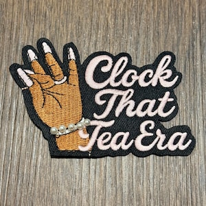 Może przedstawiać: Haftowana naszywka przedstawiająca brązową dłoń z białymi końcówkami paznokci i perłową bransoletką. Naszywka zawiera napis "Clock That Tea Era" w kursywie z różowym obrysem na czarnym tle.