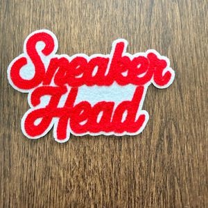 Może przedstawiać: Czerwona filcowa naszywka z napisem "Sneaker Head" w kursywie. Tekst jest obrysowany na biało i znajduje się na białym tle. Naszywka jest prawdopodobnie przeznaczona na odzież lub akcesoria.