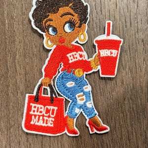 May include: Embroidered patch featuring a cartoon woman in a red sweater with "HBCU" on it, blue ripped jeans, and red heels. She carries a shopping bag that says "HBCU MADE" and a red drink cup with "HBCU" on it.