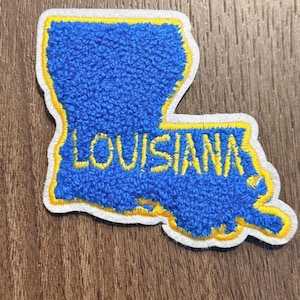 May include: A blue embroidered patch in the shape of the state of Louisiana. The patch features a textured blue surface with a yellow embroidered border and the word LOUISIANA stitched in yellow across the center, finished with a white edge.