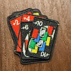 Może przedstawiać: Stos haftowanych naszywek przypominających karty do gry Uno. Naszywki mają kolory czerwony, czarny, zielony, niebieski i żółty. Karty wyświetlają liczby i symbole, z czarną obwódką i białymi szwami.