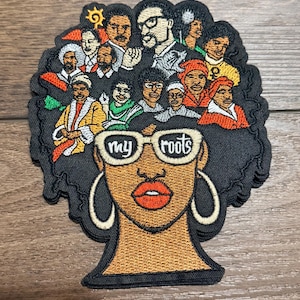 Op de afbeelding: Geborduurde patch met een zwarte vrouw met grote oorringen en een zonnebril. De zonnebril leest "my roots". Het haar van de vrouw is gevuld met portretten van historische figuren. De patch is zwart, oranje en diverse andere kleuren.