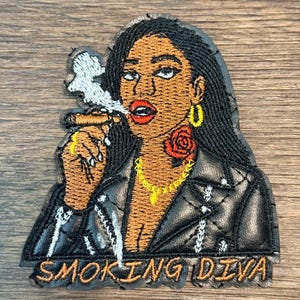 May include: Embroidered patch featuring a woman with dark hair, wearing a black leather jacket, gold jewelry, and a rose tattoo. The woman is smoking a cigar. The text "SMOKING DIVA" is at the bottom.