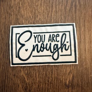 May include: A rectangular patch with the words "YOU ARE enough" in navy blue script and block letters. The patch has a white background and a navy blue border. The patch is on a wooden surface.
