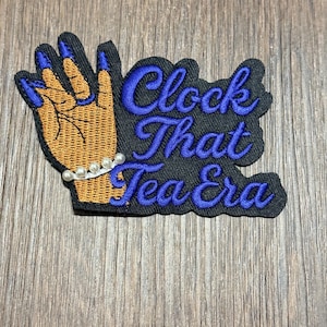 Może przedstawiać: Haftowana naszywka przedstawiająca dłoń z niebieskimi paznokciami i perłową bransoletką. Tekst "Clock That Tea Era" jest w niebieskim skrypcie na czarnym tle. Dłoń jest brązowa, a naszywka ma czarną obwódkę.