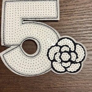 May include: A white sequined number 5 patch with a black outline and a black flower design. The patch is made of felt and sequins. The number 5 is approximately 3 inches tall.