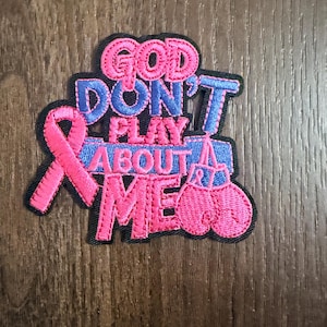 May include: An embroidered patch with a black border featuring the pink text GOD DON'T PLAY ABOUT ME. The design includes a pink awareness ribbon on the left and a pair of pink boxing gloves hanging from the letter T in the word DON'T.