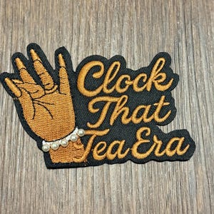 Może przedstawiać: Haftowana naszywka ze słowami "Clock That Tea Era" złotą nicią na czarnym tle. Z lewej strony znajduje się dłoń z perłową bransoletką. Naszywka znajduje się na drewnianej powierzchni.