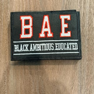Puede incluir: Parche negro rectangular con las letras "BAE" en blanco con ribete rojo. Debajo, el texto "BLACK AMBITIOUS EDUCATED" está en blanco. El parche tiene un borde negro.