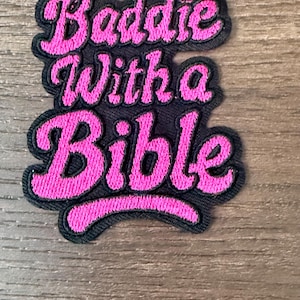 May include: Embroidered patch with the text "Baddie With a Bible" in pink script lettering on a black background. The patch has a black border and is likely for clothing or accessories.