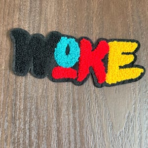 May include: A colorful patch with the word "WOKE" in a bold, playful font. The letters are in black, teal, red, and yellow, with a textured, fuzzy appearance. The patch is likely designed to be ironed on or sewn onto clothing or accessories.
