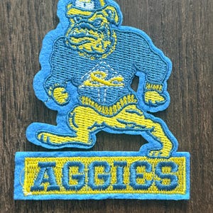May include: An embroidered patch featuring a bulldog mascot wearing a cap and sweater, coloured in shades of blue and yellow. The bulldog is depicted in a dynamic stance above a rectangular base with the text AGGIES stitched in blue on yellow.