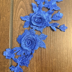 Peut inclure: Une application en dentelle florale bleu roi vif présentant deux grandes roses détaillées entourées de feuilles et de boutons plus petits. La broderie complexe est réalisée avec un motif de fil dense, créant un design texturé et en relief.