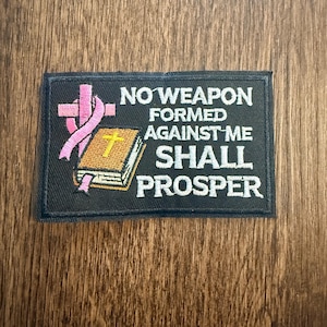 May include: Black rectangular patch with white text: "NO WEAPON FORMED AGAINST ME SHALL PROSPER." The patch features a pink ribbon wrapped around a white cross and a gold-colored book with a gold cross.