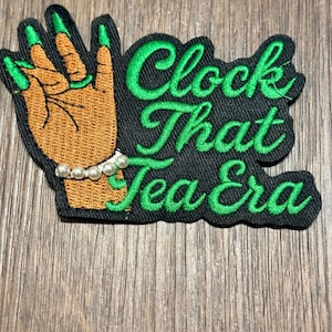 Może przedstawiać: Haftowana naszywka z napisem "Clock That Tea Era" w kolorze zielonym. Naszywka przedstawia dłoń z długimi zielonymi paznokciami i perłową bransoletką. Dłoń jest w kolorze jasnobrązowym, a naszywka ma czarne tło.