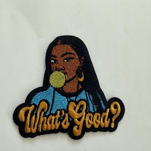 Peut inclure: Écusson brodé représentant une femme noire aux longs cheveux noirs, portant des créoles dorées et mâchant une bulle de chewing-gum verte. La phrase "What's Good?" est brodée en fil d'or.
