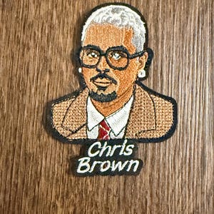 May include: Embroidered patch featuring a portrait of Chris Brown. The patch depicts a man with glasses, a beard, and a suit. The name "Chris Brown" is embroidered in white letters on a black background.