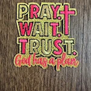May include: A gold and pink embroidered patch with the words "PRAY. WAIT. TRUST. God has a plan" in a decorative font. The patch features a pink cross and is on a wooden surface.