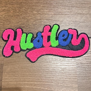 Puede incluir: Un parche colorido con la palabra "Hustler" en una fuente de escritura. Las letras son rosas, verdes y azules, con un borde negro. El parche está sobre una superficie de madera.