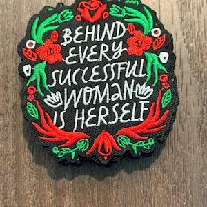 以下が含まれることがあります： 「BEHIND EVERY SUCCESSFUL WOMAN IS HERSELF」の文字が刺繍された黒いパッチ。赤、緑、白の花柄のデザイン。木製の表面に配置されています。