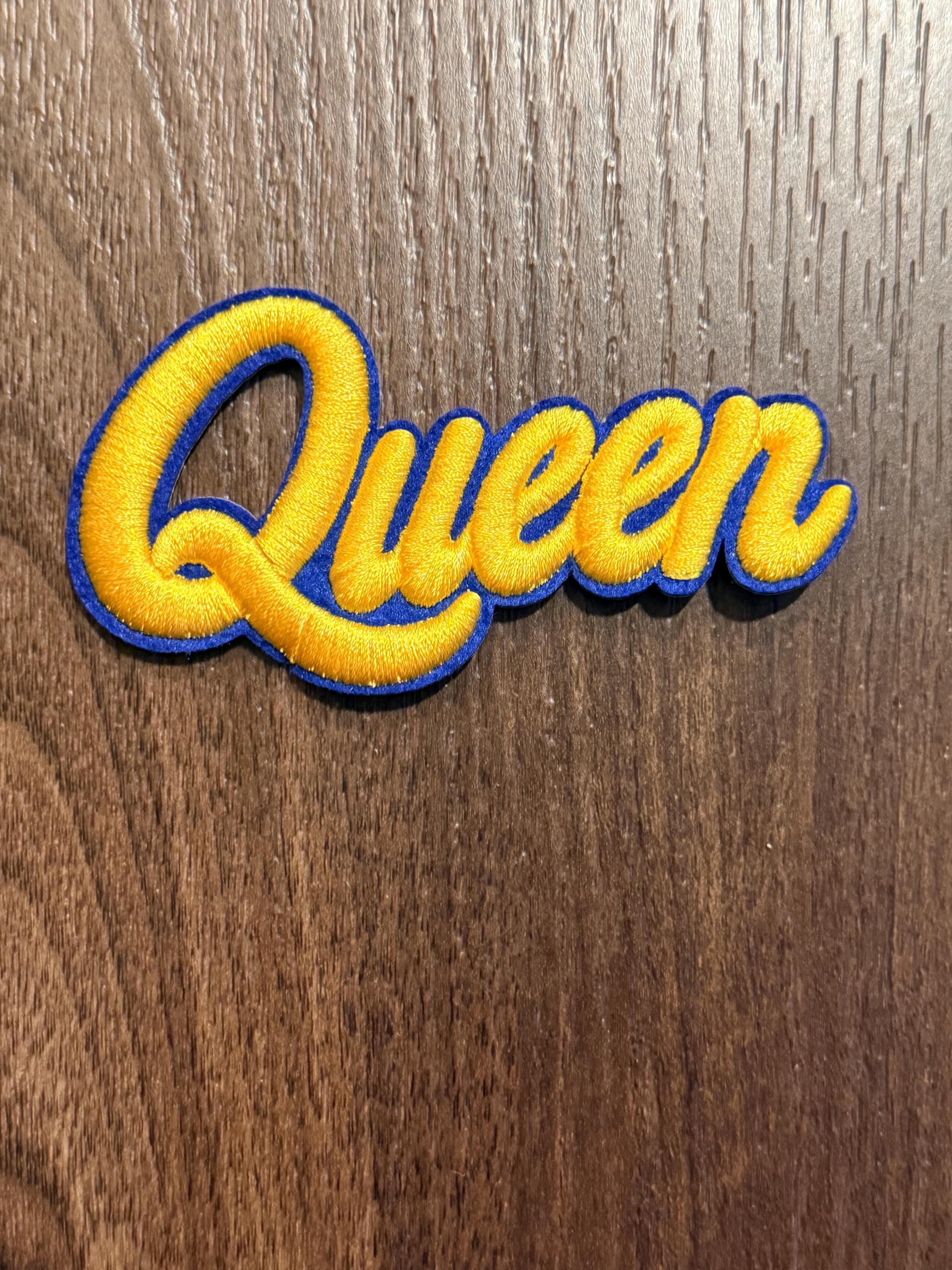 Peut inclure: Un écusson brodé représentant le mot Queen dans une police cursive. Les lettres sont jaune vif avec une bordure bleue épaisse qui suit la forme des caractères, créant un contraste marqué avec la surface en arrière-plan.