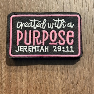 May include: A rectangular black patch with a pink border. The patch features the words "created with a PURPOSE" in white and pink lettering, with "JEREMIAH 29:11" in white at the bottom. The patch is likely an embroidered item.