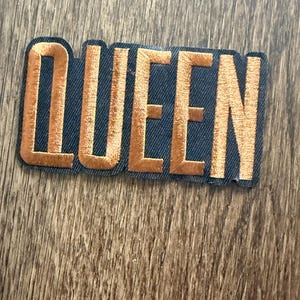 May include: Embroidered patch with the word "QUEEN" in gold thread on a navy blue background. The patch has a black border and is rectangular in shape. The patch is placed on a wooden surface.