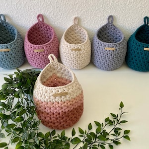 Könnte beinhalten: Sechs gehäkelte Hängekörbe in verschiedenen Farben, darunter Grau, Rosa, Beige und Türkis. Die Körbe sind mit einem strukturierten Stich gearbeitet und haben eine Schlaufe oben zum Aufhängen. Die Körbe sind in einer Reihe auf einer weißen Oberfläche angeordnet.