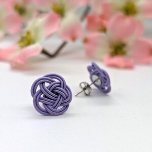 May include: A pair of lavender stud earrings with a woven knot design. The earrings are set against a white background, with blurred pink flowers in the background. The earrings are a delicate accessory.