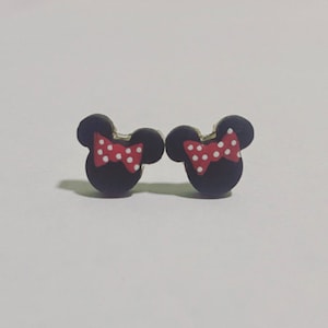 May include: A pair of black stud earrings in the shape of a mouse head, each with a red bow featuring white polka dots. The earrings are small and likely made of a lightweight material.