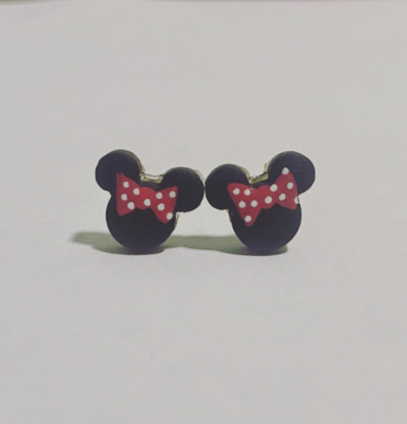 May include: A pair of black stud earrings in the shape of a mouse head, each with a red bow featuring white polka dots. The earrings are small and likely made of a lightweight material.
