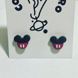 May include: A pair of black and red Mickey Mouse-shaped stud earrings. The earrings feature a black head with two round ears and a red face with two white ovals. The earrings are presented on a white card.