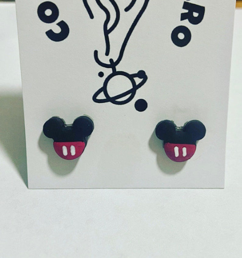 May include: A pair of black and red Mickey Mouse-shaped stud earrings. The earrings feature a black head with two round ears and a red face with two white ovals. The earrings are presented on a white card.