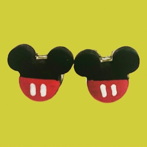 May include: A pair of Mickey Mouse earrings. Each earring has a black head with two round ears and a red bottom with two white vertical stripes. The earrings are set against a yellow background.